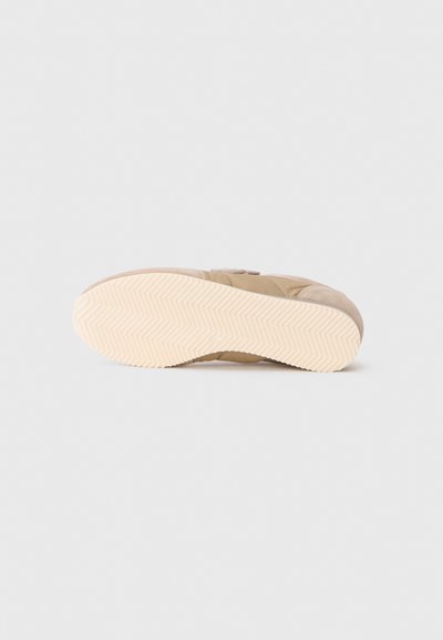 Beige sneaker sedd från sulan, som visar en ljusgummisula med ett fiskbensmönster på vit bakgrund.