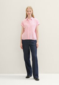Woman wearing a pink and white vertical striped short-sleeve blouse, dark blue wide-leg pants, and black shoes, standing against a plain background.