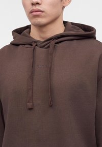 Homme portant un sweat à capuche marron avec capuche et cordons, avec le mot "HUGO" embossé sur la poitrine.