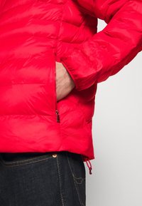 Röd puffjacka med kviltat mönster, en sidoficka med dragkedja, elastiska ärmslut och snörning i nederkant. Slät, glänsande tygtextur.