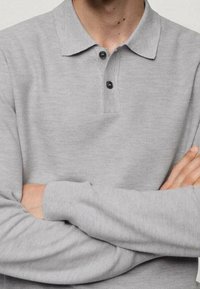 Mann trägt ein hellgraues Langarm-Polohemd mit zwei schwarzen Knöpfen, die Arme vor der Brust verschränkt.