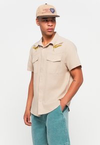 Light beige short-sleeve shirt with two front pockets, yellow embroidered detailing on shoulders, paired with teal denim shorts and a beige cap.