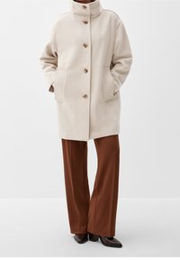 Beige vlnený kabát s vysokým límcem, zapínáním na knoflíky v přední části a bočními kapsami, zkombinovaný s hnědými širokými kalhotami a tmavými botami.
