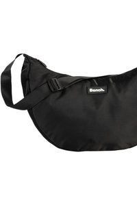 Zwarte nylon schoudertas met een gebogen vorm, verstelbare schouderband en een rechthoekig logopatch. Glad oppervlak met een ritssluiting.