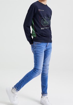 Osoba nosící tmavě modrou mikinu se zelenými bočními panely a diagramem jízdního kola, světle modré džíny, bílé ponožky a bílé tenisky.