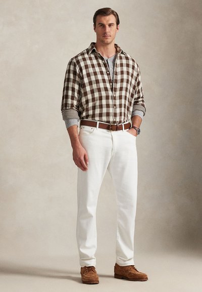 Camisa xadrez castanha e bege, jeans brancos, cinto de couro castanho e sapatos castanhos. Camiseta cinza visível por baixo da camisa.