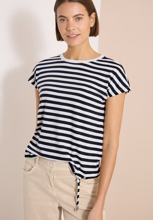 Vrouw draagt een zwart-wit gestreepte top met korte mouwen en een zijknoop, gecombineerd met lichtbeige broek tegen een neutrale achtergrond.
