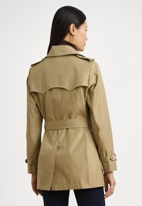 Trenci kaki cu o croială ajustată, talie cu curea și un detaliu mare la spate. Prezintă mâneci croite cu manșete reglabile.