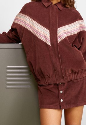 Kvinde iført en brun manchetjakke med lyserøde og hvide chevronstriber og matchende nederdel med sølv trykknapper.