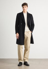 Cappotto doppio petto nero in tessuto texture, indossato sopra un maglione a coste crema, abbinato a pantaloni beige e scarpe nere.