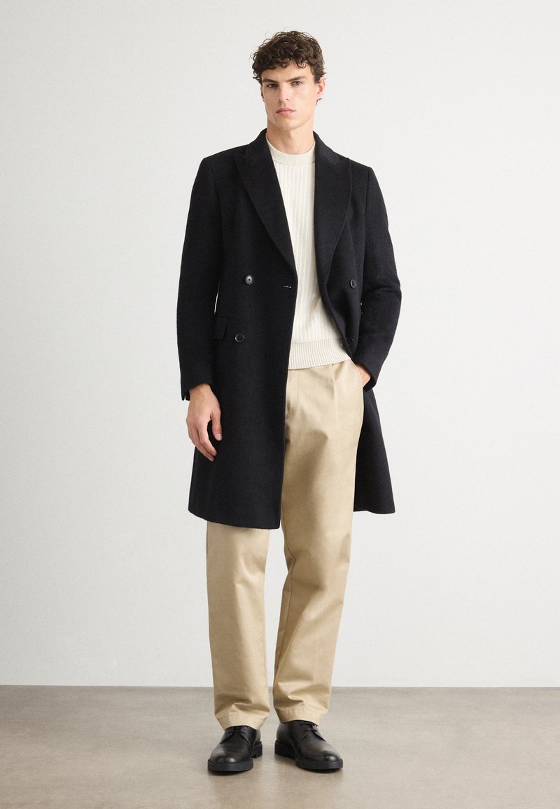 Cappotto doppio petto nero in tessuto texture, indossato sopra un maglione a coste crema, abbinato a pantaloni beige e scarpe nere.