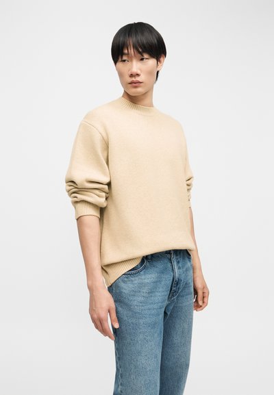 Jeune personne portant un pull beige à manches longues retroussées et un jean bleu, debout devant un fond clair uni.