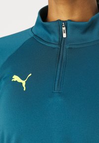Türkisfarbenes Sportshirt mit halbem Reißverschlusskragen, aus strukturiertem Stoff. Mit kleinem gelben Puma-Logo auf der Brust.