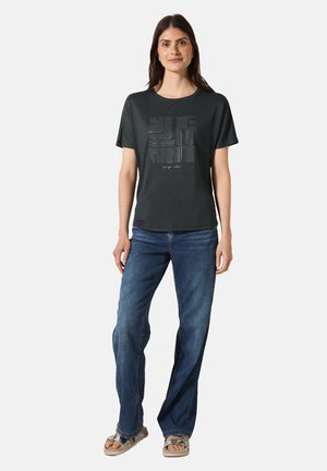 T-shirt gris foncé à manches courtes avec un design graphique sur le devant, associé à un jean en denim bleu et des sandales de couleur claire.