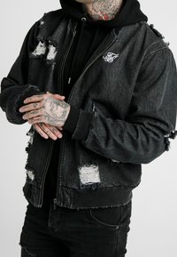 Czarna kurtka bomber z denimu z przetarciami, posiadająca postrzępione krawędzie i podarte łatki. Wyposażona w zamek błyskawiczny oraz haftowane logo.