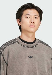 Grå sweatshirt med ribbad halsringning och texturerade horisontella ränder längs axlarna. Litet svart logo på bröstet. Mjukt material.