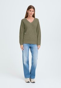 Olijfgroene gebreide trui met een V-hals, lange mouwen en een textuurstof, gecombineerd met lichtblauwe rechte jeans en beige sneakers.