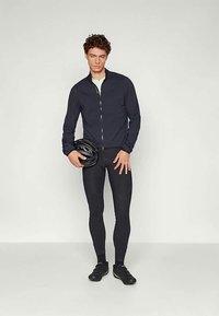 Ung mann iført svart sykkelantrekk og holder en svart sykkelhjelm, stående mot en lys, ensfarget bakgrunn.