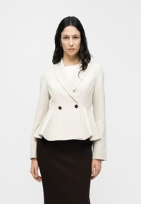 Blazer crème à basque avec un design croisé, deux boutons noirs et des épaules structurées. Associé à une jupe marron côtelée.