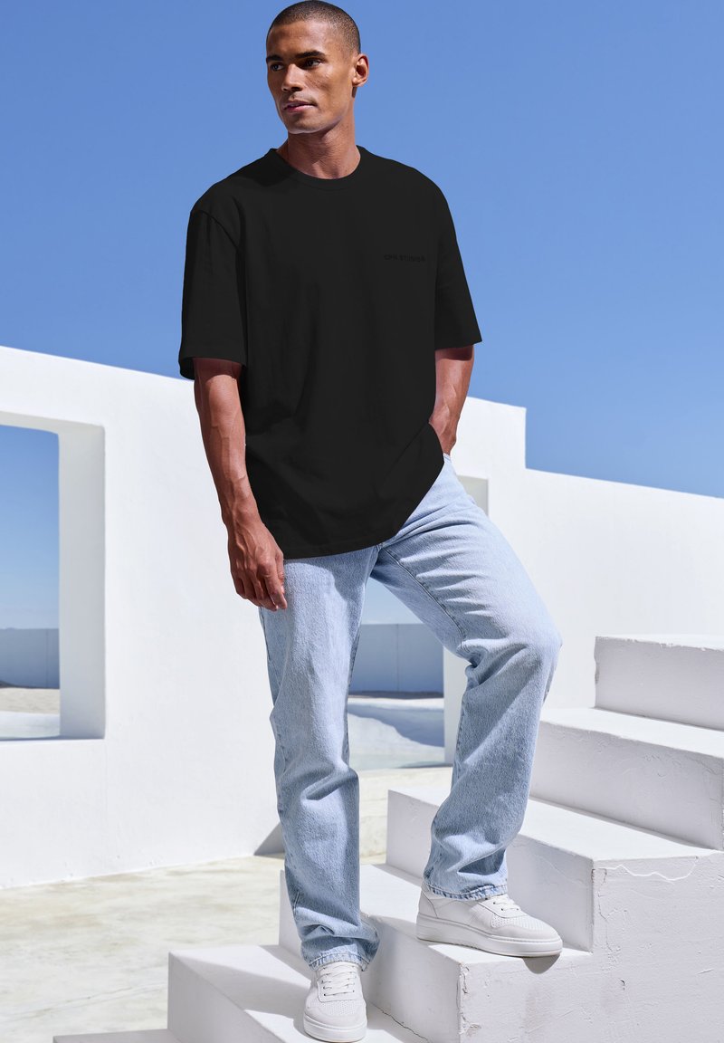 Zwarte T-shirt met korte mouwen en een minimalistisch ontwerp, gecombineerd met lichtblauwe jeans en witte sneakers, staand op witte trappen tegen een blauwe lucht.