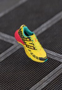 Zapatilla de correr amarilla brillante con acentos negros, parte superior de malla con patrón, cuello turquesa y talón rojo, con una suela texturizada para mayor agarre.