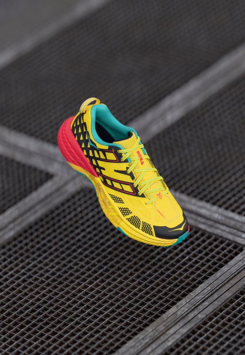 Zapatilla de correr amarilla brillante con acentos negros, parte superior de malla con patrón, cuello turquesa y talón rojo, con una suela texturizada para mayor agarre.