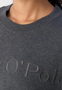 Gros plan d'une personne portant un sweat-shirt gris foncé avec le texte en relief « POLO » sur la poitrine.