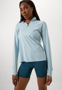 Light blue long-sleeve athletic top with a zippered collar, paired with dark teal shorts. Fabric appears smooth and stretchy, with a fitted design.