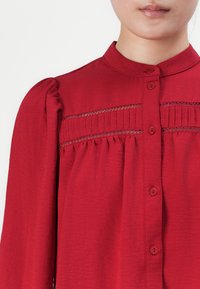 Kvinne iført rød langermet bluse med folder, blonder og rund krage, knappet foran.