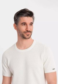 Bílý texturovaný pletený svetr s krátkým rukávem a kulatým výstřihem, se subtilním kovovým akcentem na rukávu. Hladký povrch látky.