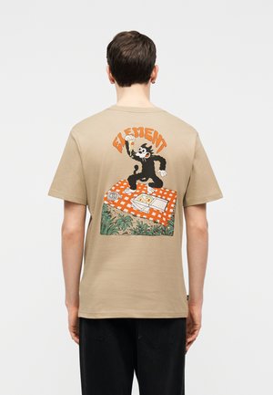 Personne portant un t-shirt beige avec un dessin de chat noir tenant une tranche de pizza, assise au-dessus d'une couverture de pique-nique à carreaux orange et entourée de plantes en dessous.