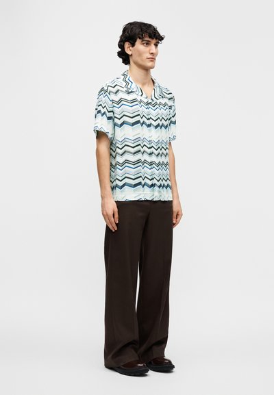 Κοντομάνικο πουκάμισο με ζιγκ-ζαγκ μοτίβο σε τιρκουάζ και λευκό, κατασκευασμένο από ελαφρύ ύφασμα. Συνδυασμένο με χαλαρά καφέ παντελόνια και σκούρα παπούτσια.