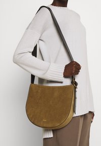 Brun mocka crossbody-väska med en böjd form, svart läderrem och subtil gravyr av varumärket på framsidan. Mjuk textur och minimalistisk design.