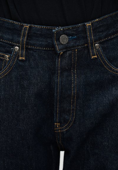 Κοντινή λήψη σκούρου μπλε τζιν με πορτοκαλί ραφές, μεταλλικό κουμπί κλεισίματος και θηλιές για ζώνη πάνω σε μαύρο ύφασμα.