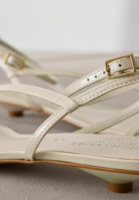 Gros plan sur des sandales crème à lanières, petits talons et détails de boucles dorées sur une surface neutre.