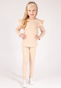 Beżowa prążkowana bawełniana bluzka z długim rękawem i falbankami na rękawach, dopasowane prążkowane legginsy oraz opaska na głowę z węzłem. Białe sneakersy z akcentami.