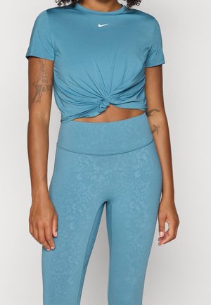 Woman wearing light blue twisted knot crop top with a white Nike logo and high-waisted textured floral leggings, showing arm tattoos.