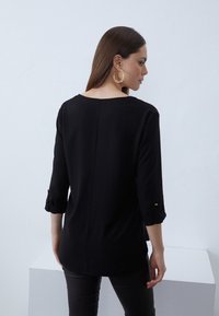 Black blouse with three-quarter sleeves, a low scoop neck, and roll-up cuffs. Smooth fabric with a straight cut and back seam detail.