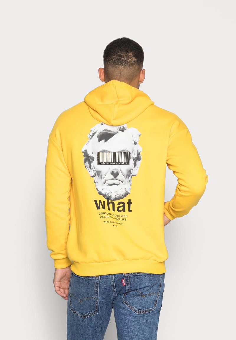 Żółta bluza z kapturem z grafiką rzeźbionej twarzy z kodem kreskowym na oczach. Dodatkowy tekst poniżej zawiera słowa "co" oraz "zjedz swój umysł".