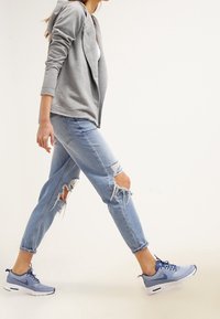 Un sweat à capuche gris zippé, un haut blanc, un jean bleu clair avec des déchirures, et des baskets bleues Nike avec des semelles blanches sont visibles.