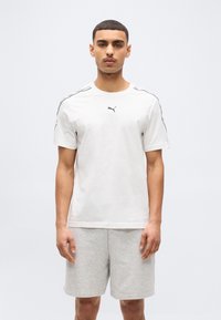 Νέος άνδρας που φοράει λευκό μπλουζάκι Puma με μαύρο λογότυπο και ανοιχτό γκρι σορτς, στέκεται μπροστά σε απλό λευκό φόντο.