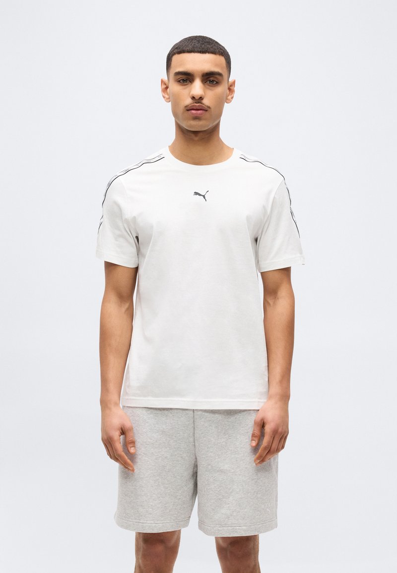 Νέος άνδρας που φοράει λευκό μπλουζάκι Puma με μαύρο λογότυπο και ανοιχτό γκρι σορτς, στέκεται μπροστά σε απλό λευκό φόντο.