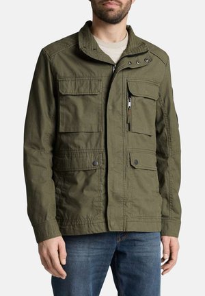 Homme portant une veste utilitaire vert olive avec plusieurs poches à l'avant et un jean bleu, debout devant un fond blanc uni.
