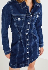 Denimová šaty v tmavě modré barvě s předním zapínáním na knoflíky, límečkem a dlouhými rukávy. Mají kapsy na hrudi a přiléhavý střih s viditelným švem.