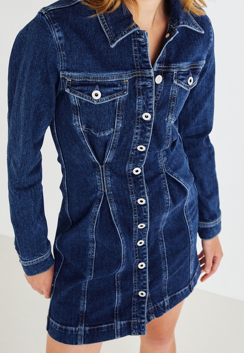 Denimová šaty v tmavě modré barvě s předním zapínáním na knoflíky, límečkem a dlouhými rukávy. Mají kapsy na hrudi a přiléhavý střih s viditelným švem.