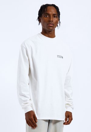 Man met gevlochten haar die een wit shirt met lange mouwen draagt met kleine zwarte tekst op de linkerborst, staand tegen een effen witte achtergrond.