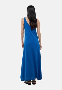 Robe maxi sans manches bleue avec un dos nu, ayant une coupe décontractée et une texture douce. Portée avec des sandales noires.