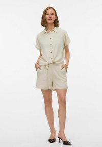 Beige linnen korte romper met een gekraagde shirt, knopen aan de voorkant, korte mouwen, een trekkoord in de taille en zijzakken. Gemaakt met donkere hakken.