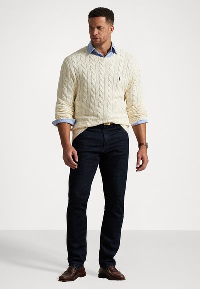 Suéter de punto trenzado crema, camisa de color azul claro debajo, jeans oscuros, zapatos marrones. Diseño ajustado, puños acanalados, detalle sutil del logo.