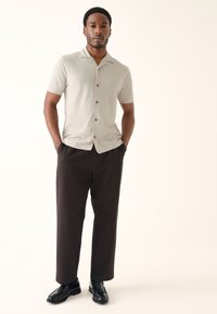 Ljusgrå kortärmad skjorta med notch-krage, mörkbruna veckade byxor och svarta skor; slät textur och knappdetaljer.